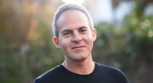 Portrait of a middle-aged man with short gray hair wearing a black shirt, smiling outdoors with blurred greenery in the background.